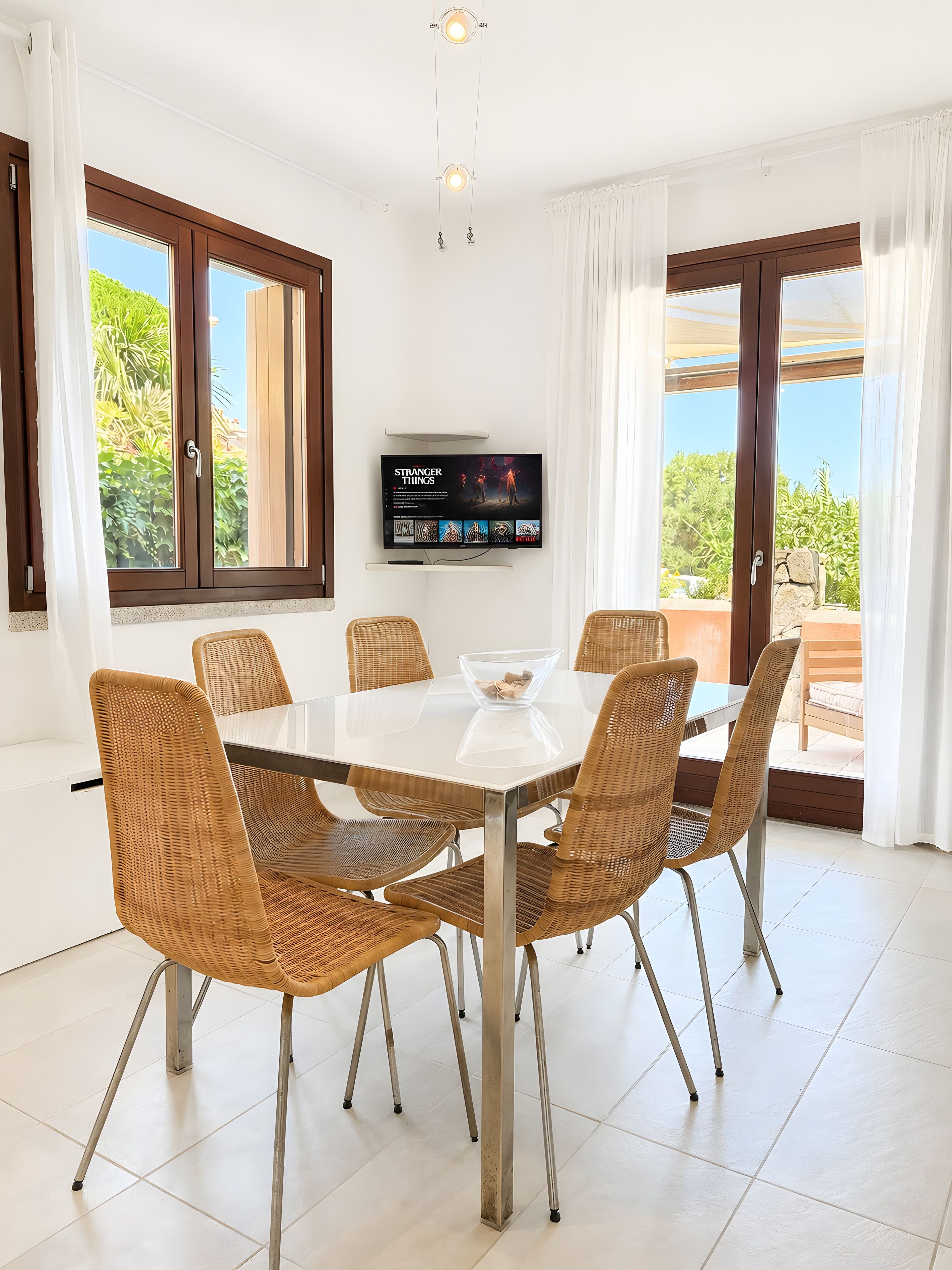 A dining area with a glass table and wicker chairs, a TV on the wall, and large windows with curtains.