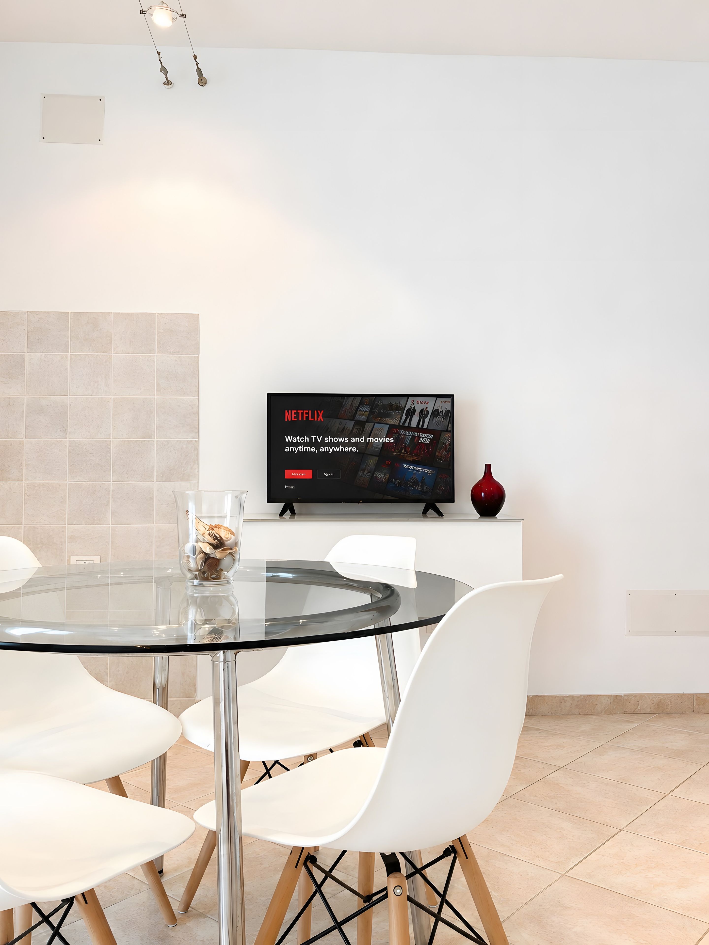 Un espace repas moderne avec une table en verre, des chaises blanches, un téléviseur à écran plat fixé au mur et un vase posé sur un meuble.