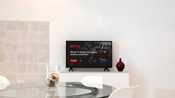 Un espace repas moderne avec une table en verre, des chaises blanches, un téléviseur à écran plat fixé au mur et un vase posé sur un meuble.