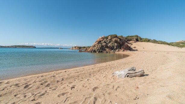 Una spiaggia sabbiosa con acque limpide, una grande formazione rocciosa e una vista sullo sfondo del mare.