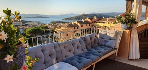 A balcony with a cushioned bench, potted plants, and a view of coastal buildings and mountains.