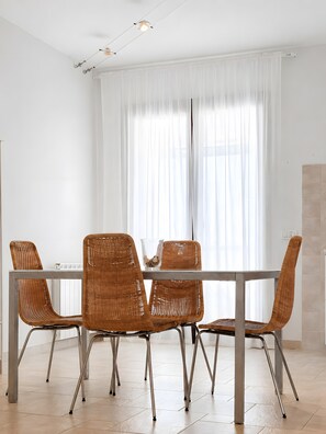 Un coin repas avec une table et quatre chaises en osier, un vase en verre et un rideau blanc.