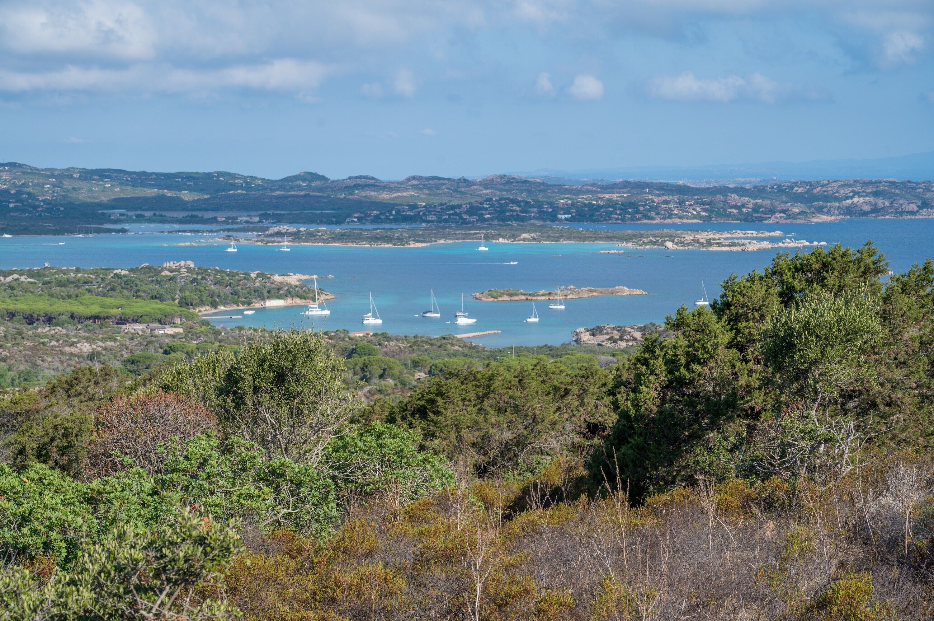 Un paysage côtier avec des bateaux, de la végétation et des collines à l’horizon.