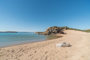 A sandy beach with clear water, a large rock formation, and a distant view of the sea.