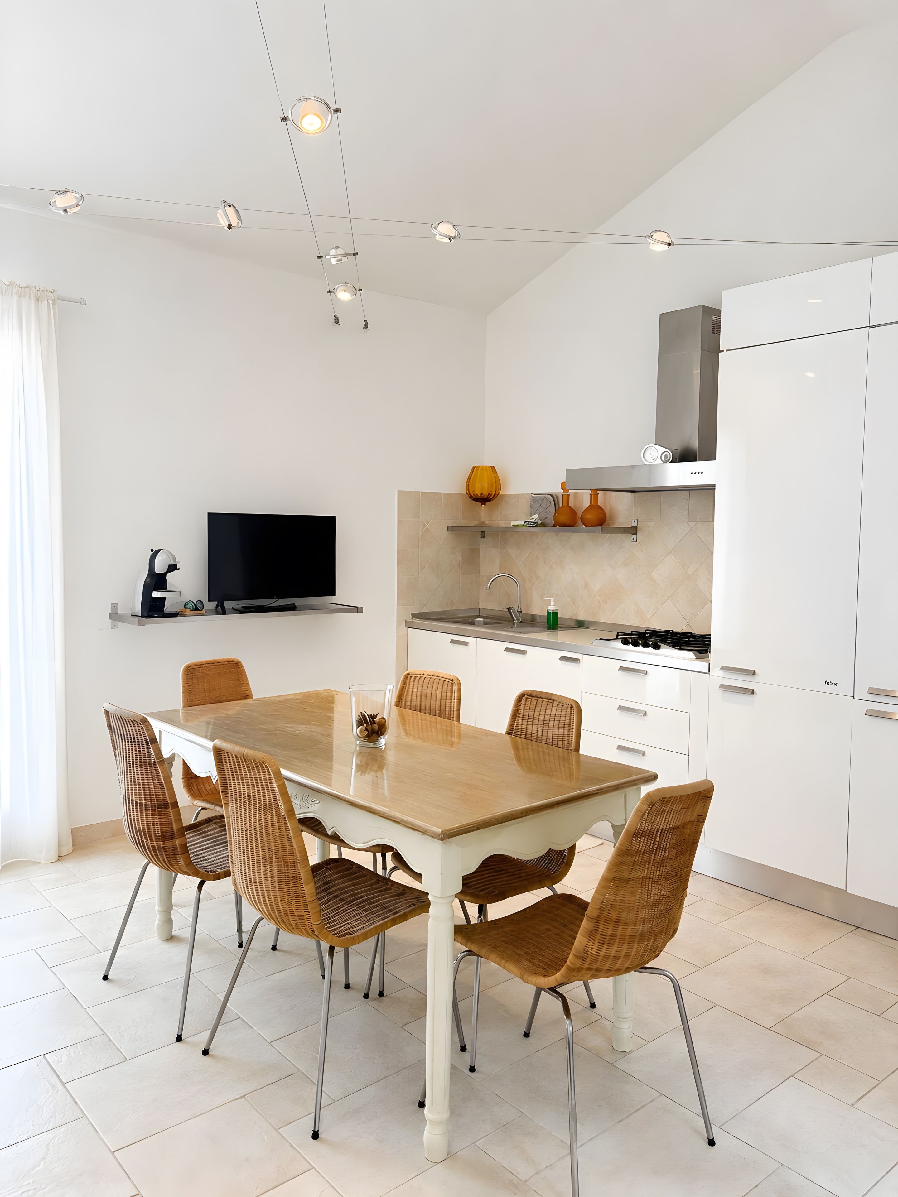 Una cucina moderna con mobili bianchi, un tavolo da pranzo con sedie in vimini e un televisore a schermo piatto su uno scaffale.