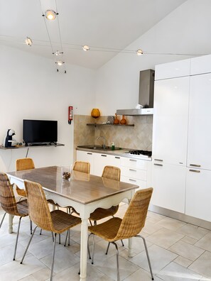 Une cuisine moderne avec des meubles blancs, une table à manger entourée de chaises en osier et un téléviseur à écran plat.