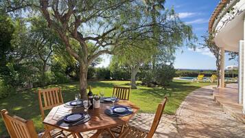 A wooden outdoor table set for dining with chairs, a pool in the background, and a clear sky.