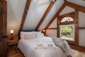 Un dormitorio con una cama grande, vigas de madera, un ventanal con vistas y una mesita de noche con una lámpara.