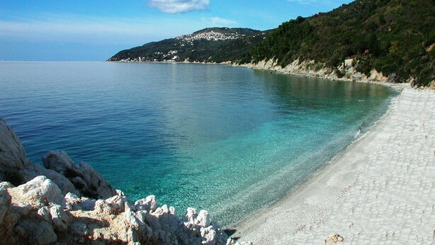 A coastal beach with clear turquoise water, rocky shoreline, and a hilly backdrop with buildings. - Cozy Traditional Home with Magnificent Sea Views & Garden (Skopelos)