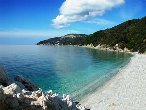 Ein Küstenstrand mit klarem türkisblauem Wasser, felsigem Ufer und einer hügeligen Landschaft mit Gebäuden im Hintergrund.