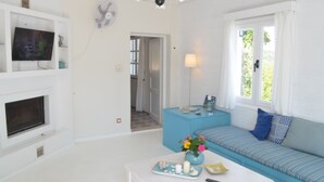 A living room with a TV, a fan, a sofa, a coffee table, and a window with curtains. - Cozy Traditional Home with Magnificent Sea Views & Garden (Skopelos)