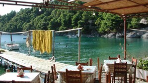 An outdoor dining area with tables and chairs, a wooden dock extending into a lake, and a backdrop of green hills. - Cozy Traditional Home with Magnificent Sea Views & Garden (Skopelos)