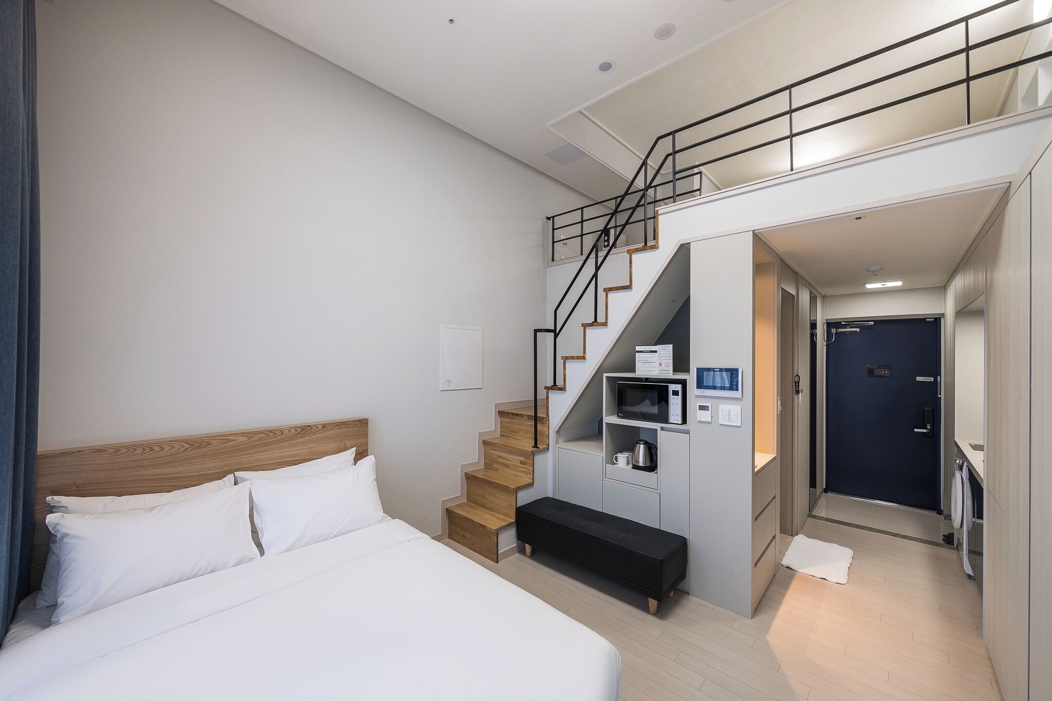 A modern hotel room with a wooden staircase, a bed with white linens, a small bench, and a microwave on a shelf.