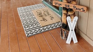 Varanda de madeira com tapete de entrada que diz "Entre e Saia", um urso de pelúcia com gorro de Papai Noel e uma placa que diz "Limpe as Patas".