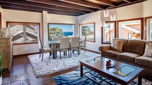 A spacious living room with a wooden ceiling, large windows, a dining area, and a cozy seating area. - Seafarer - Pacific Grove Beach House - Steps to the Water, Incredible Ocean Views (Pacific Grove)