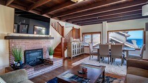 A spacious living room with a brick fireplace, wooden ceiling beams, a dining area with a view of the ocean, and a staircase leading to an upper level. - Seafarer - Pacific Grove Beach House - Steps to the Water, Incredible Ocean Views (Pacific Grove)