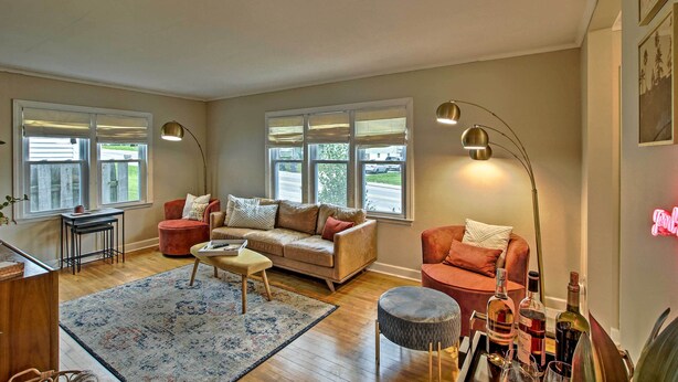 A spacious living room with a beige sofa, two armchairs, a coffee table, and a rug. There are two windows with blinds, a floor lamp, and a neon sign on the wall. - Ideal Green Bay Home < 3 Mi to Lambeau Field! (Green Bay)