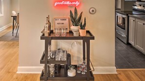 A kitchen with a wooden floor, a black metal cart with wine glasses and bottles, a neon sign, and a potted plant. - Ideal Green Bay Home < 3 Mi to Lambeau Field! (Green Bay)