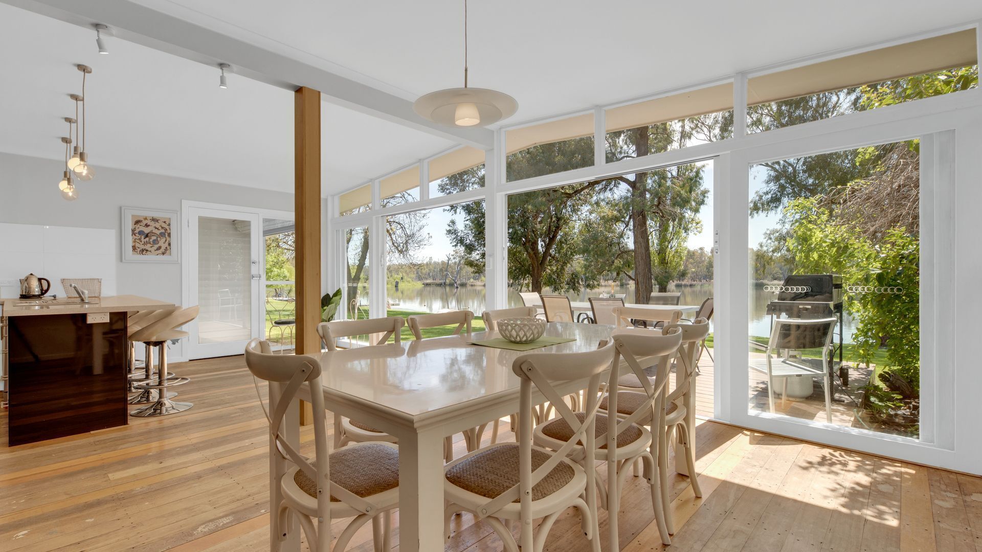 Un vaste espace à manger avec une grande table et des chaises, une cuisine dotée d’un plan de travail en bois et de grandes fenêtres offrant une vue sur un lac et des arbres.
