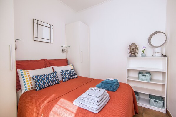 A neatly made bed with an orange bedspread, a white headboard, and a variety of pillows. There is a white shelf with storage boxes and a decorative Buddha statue. - Co-ownership (Nice)