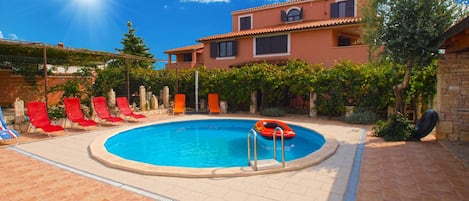 A swimming pool with red lounge chairs, a lifebuoy, and a stone building in the background.