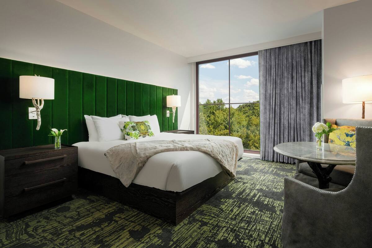 A hotel room with a large bed, green headboard, two bedside tables with lamps, a round table with a chair, and a view of greenery outside.