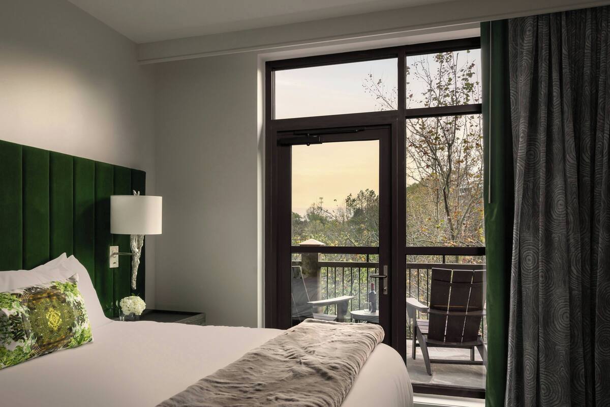 A hotel room with a large bed, a green headboard, a nightstand with a lamp, and a view of the outdoors through a large window.