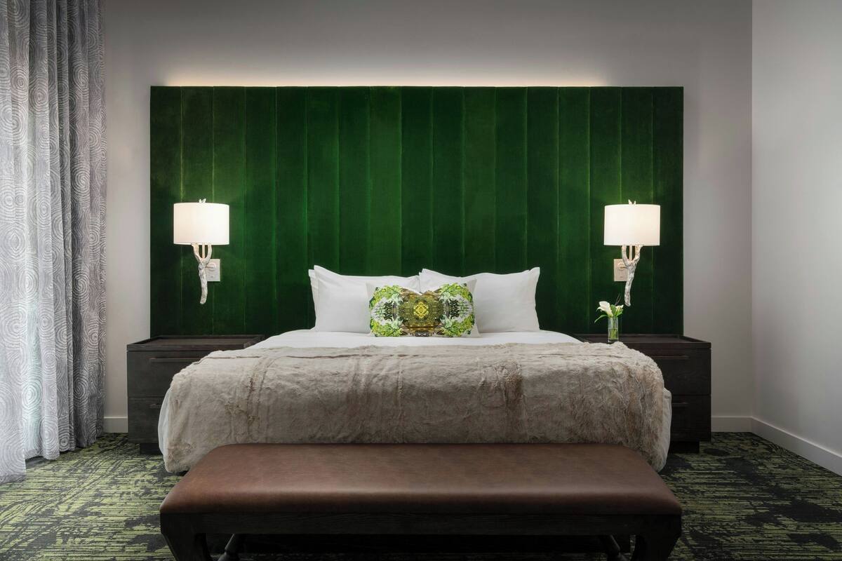 A hotel room with a large bed, two bedside lamps, a bench, and a green headboard.