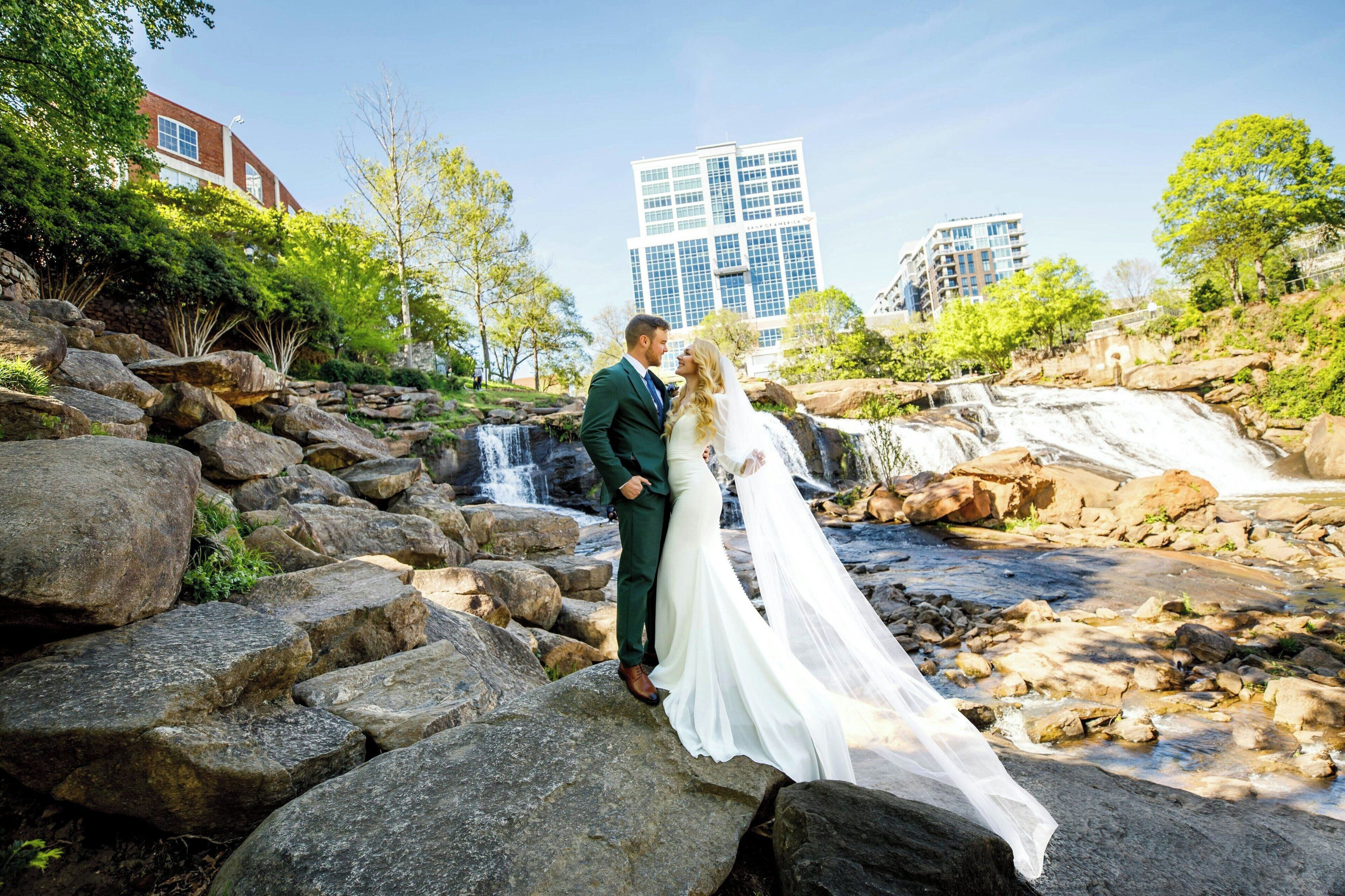 A couple standing on rocks near a waterfall with buildings in the background.