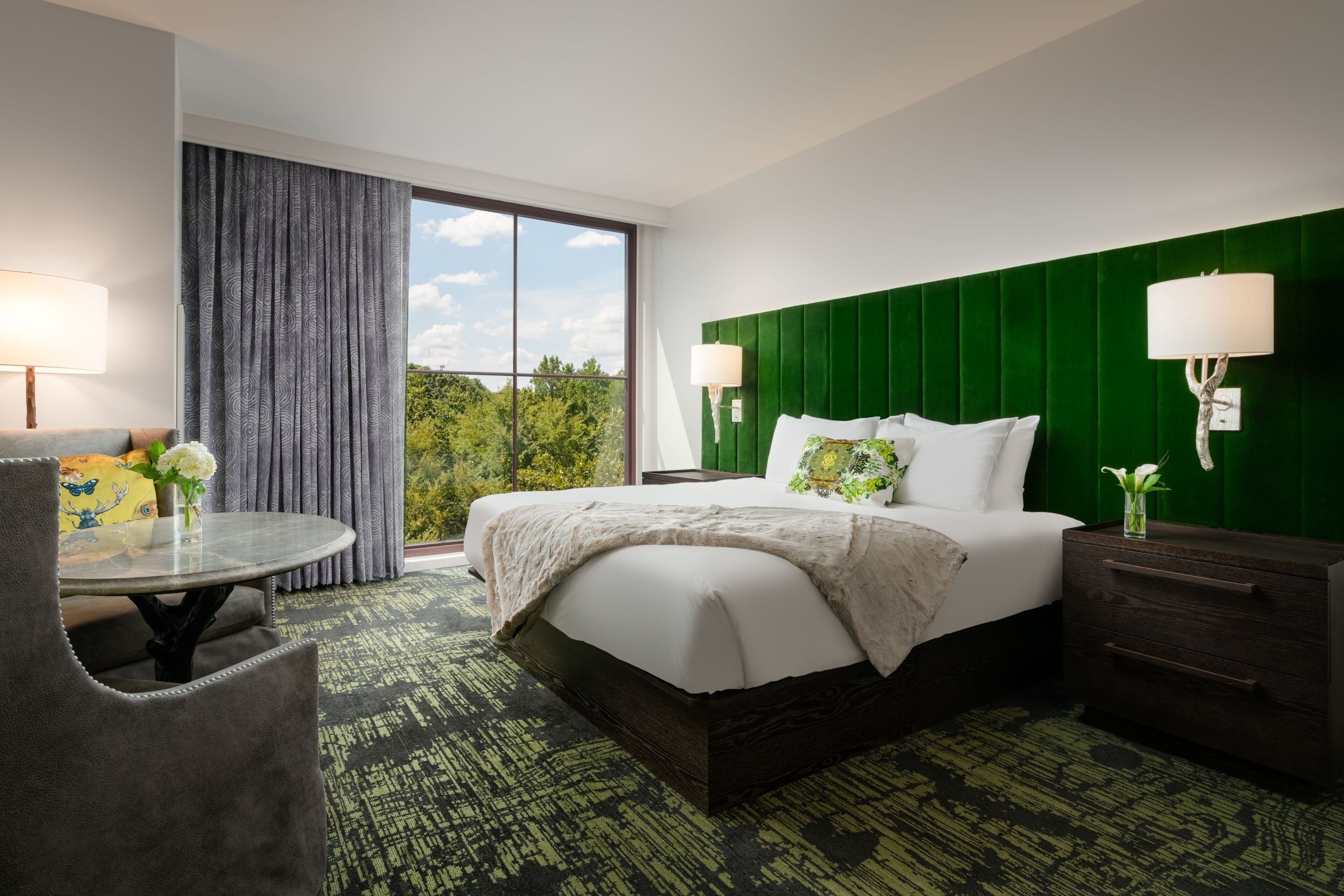 A hotel room with a large bed, a green headboard, a window with curtains, a small table with a lamp, and a chair.