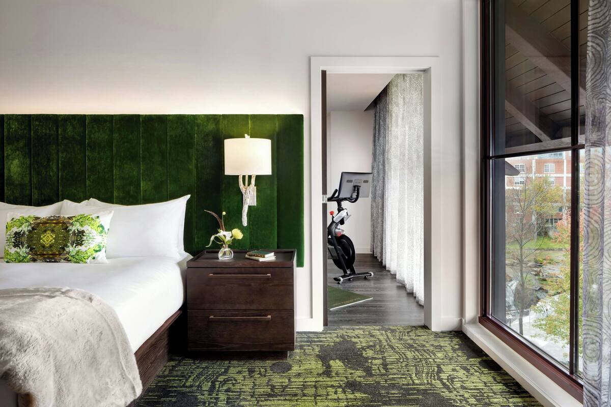 A hotel room with a large bed, a nightstand, a lamp, a view of a bike outside, and a window with a view of buildings.