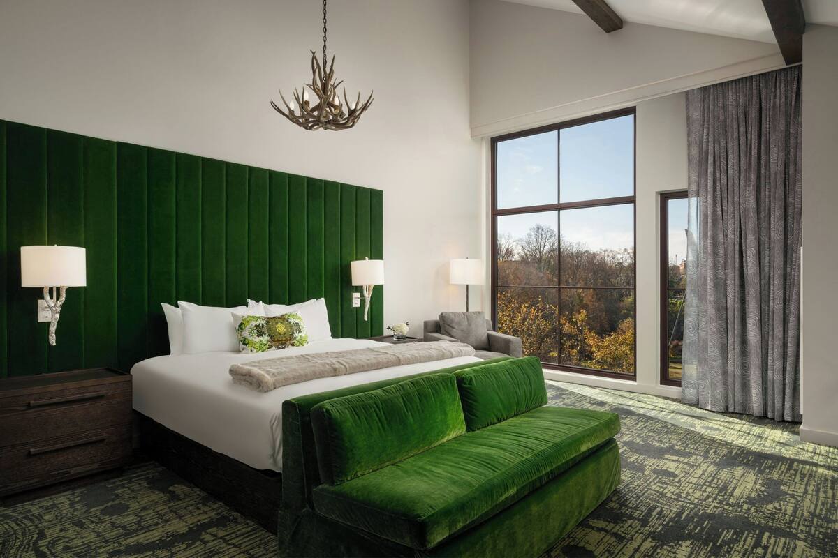 A modern hotel room with a large bed, green upholstered headboard, two bedside lamps, a green sofa, and a large window with a view of trees.