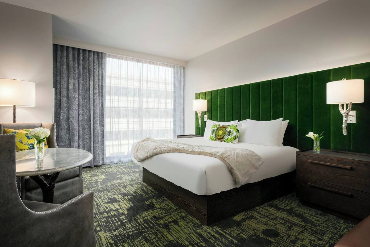 A hotel room with a bed, bedside lamps, a small table with a chair, and a large window with curtains.