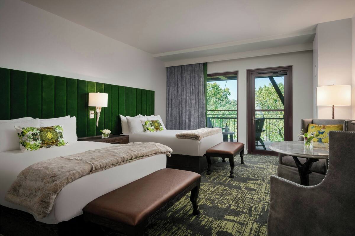 A hotel room with two beds, a bench, a table, and a balcony with a view.