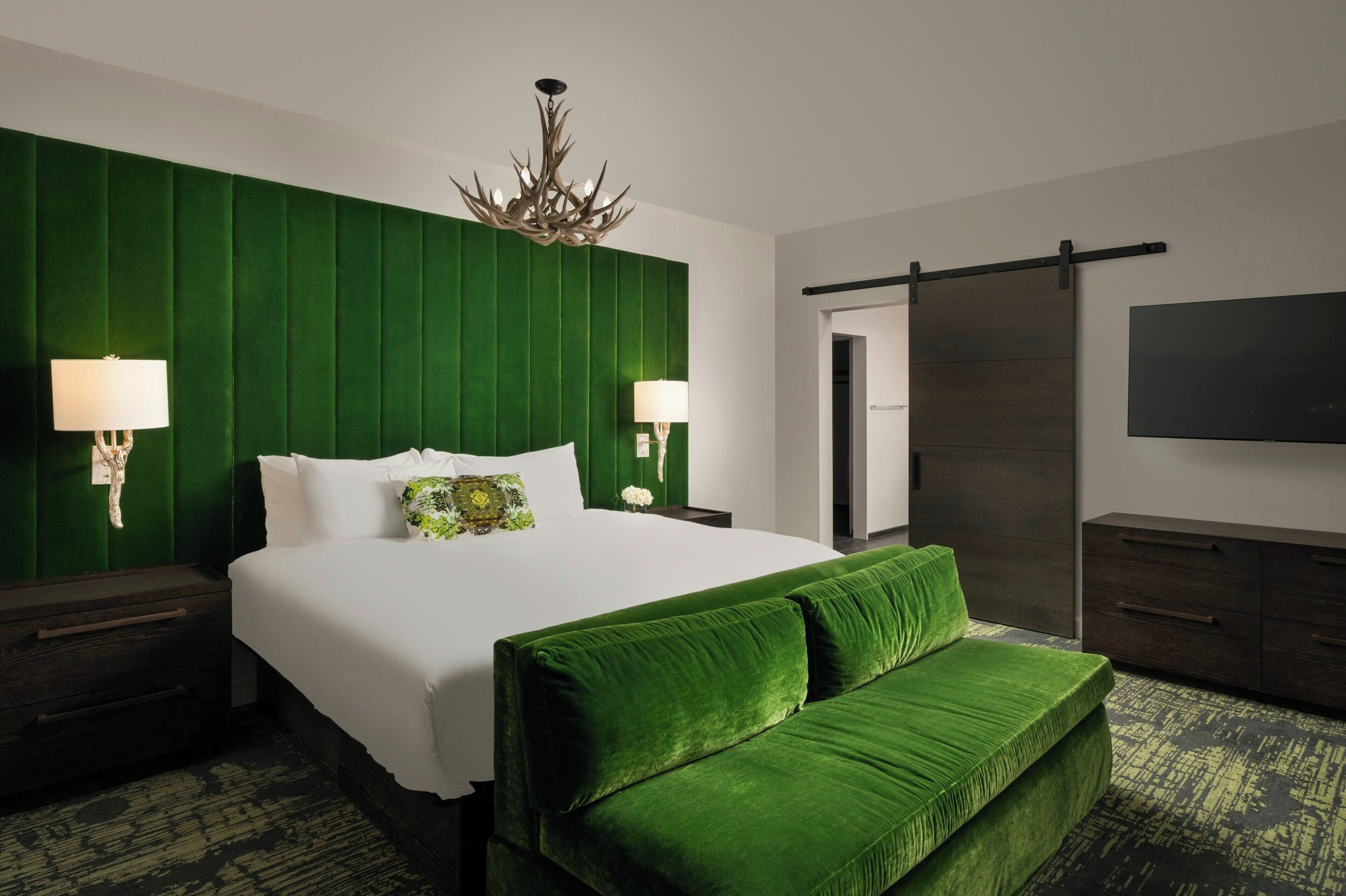 Habitación de hotel con una cama grande, cabecera tapizada de verde, dos lámparas de noche, un candelabro, un armario de madera oscura y un televisor de pantalla plana en la pared.