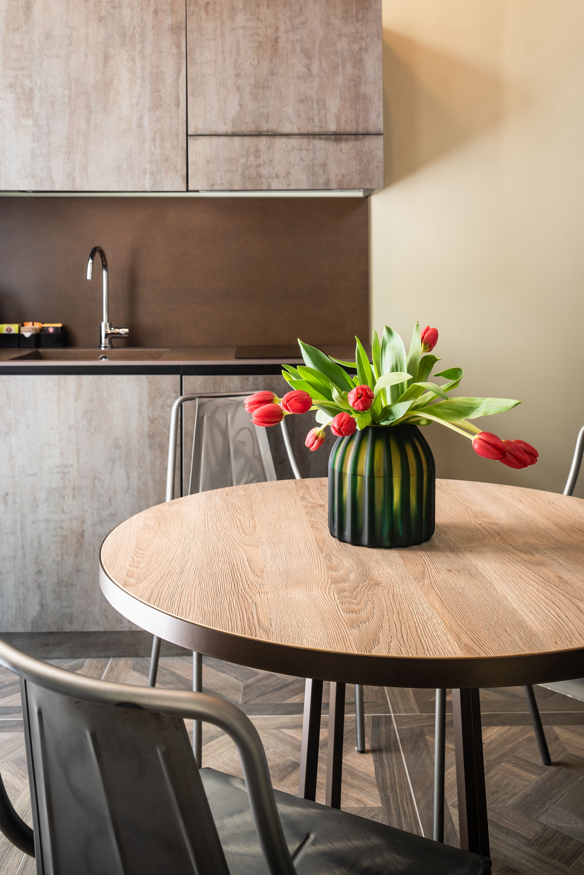 A round wooden table with a vase of red tulips in the center, surrounded by metal chairs.