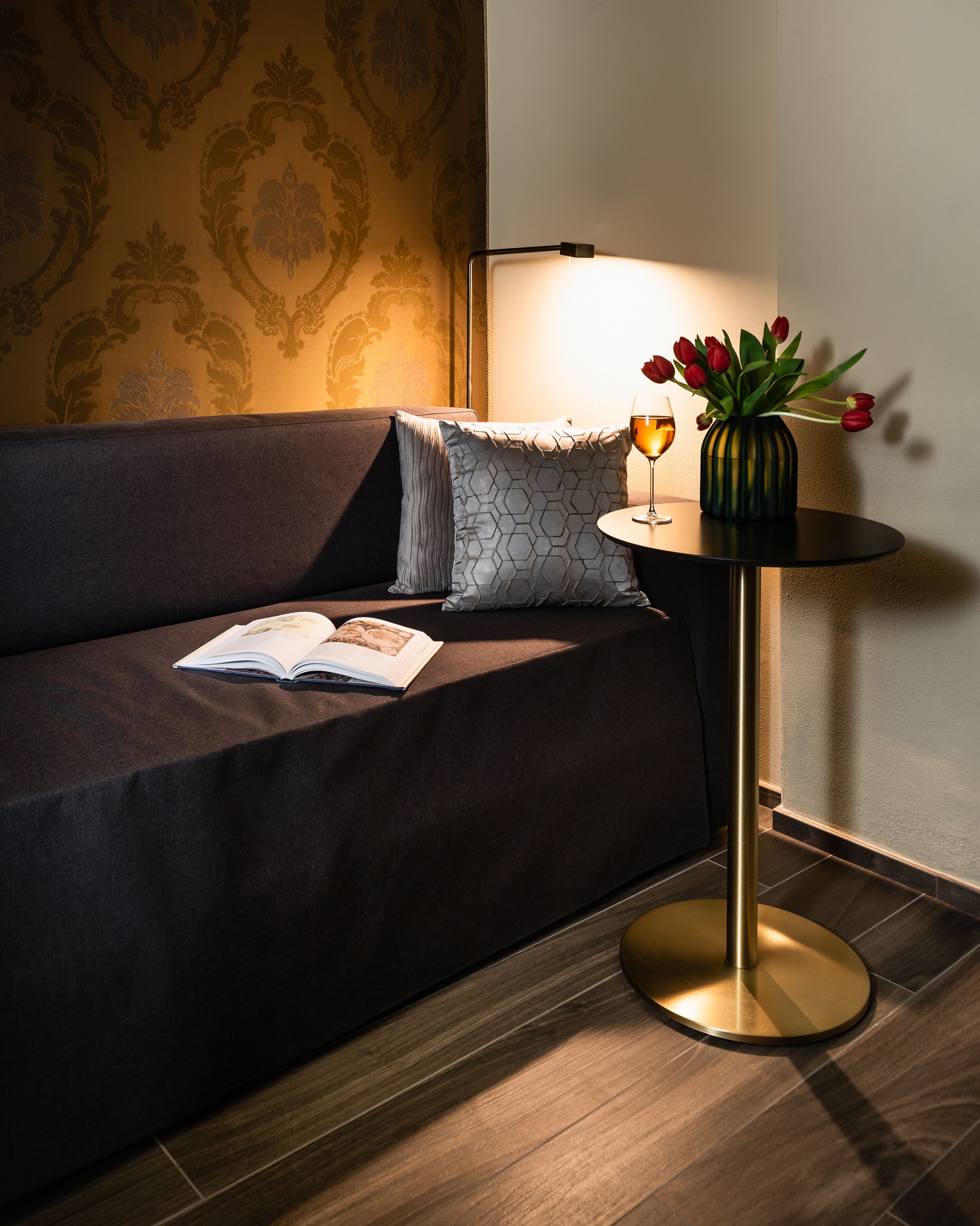 A modern living room with a dark sofa, a round side table with a lamp, a vase of red tulips, and a book on the sofa.