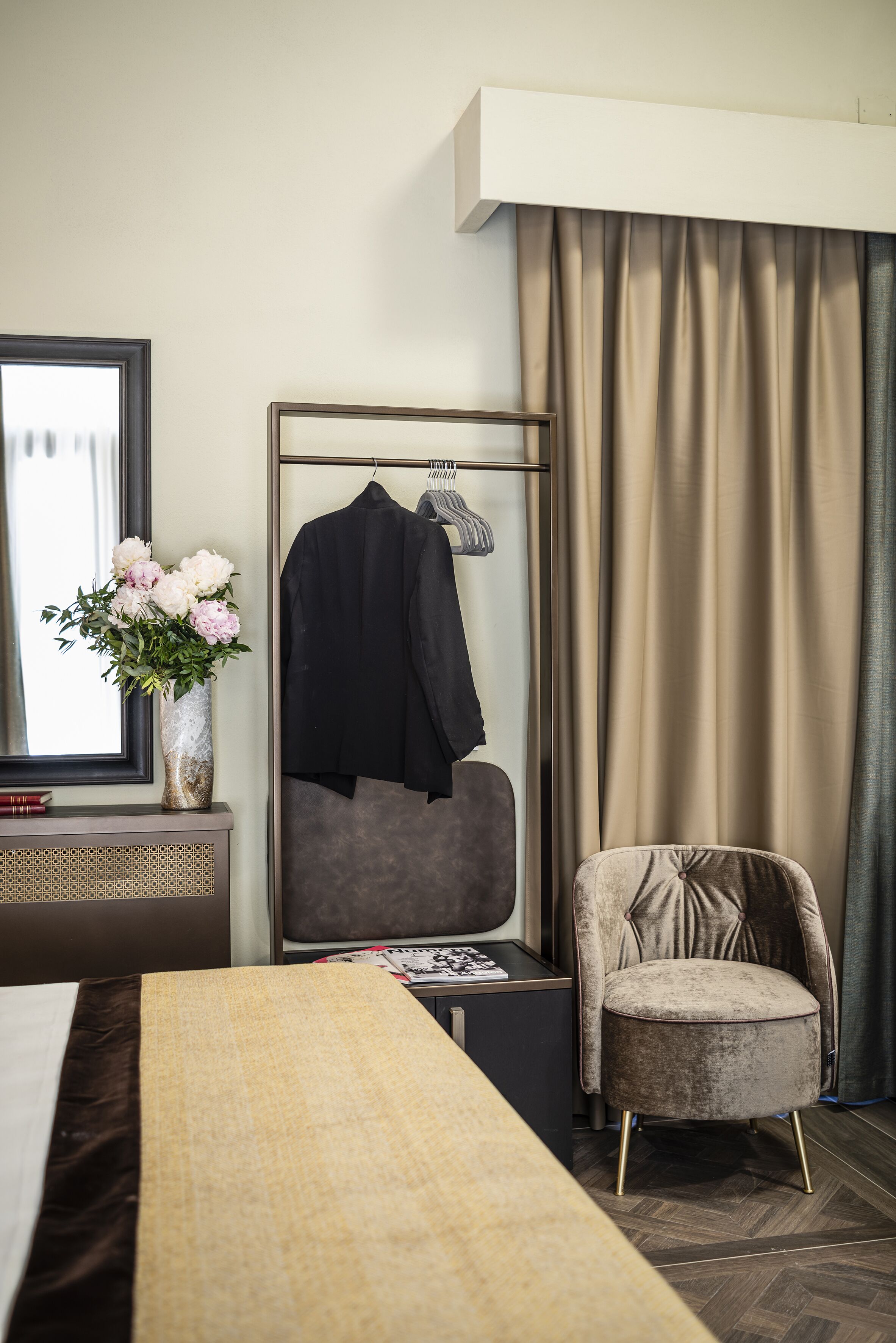 Ein Hotelzimmer mit einem Bett, einem Sessel, einem Spiegel, einer Kommode, einem Kleiderhaken und einem Fenster mit Vorhängen.