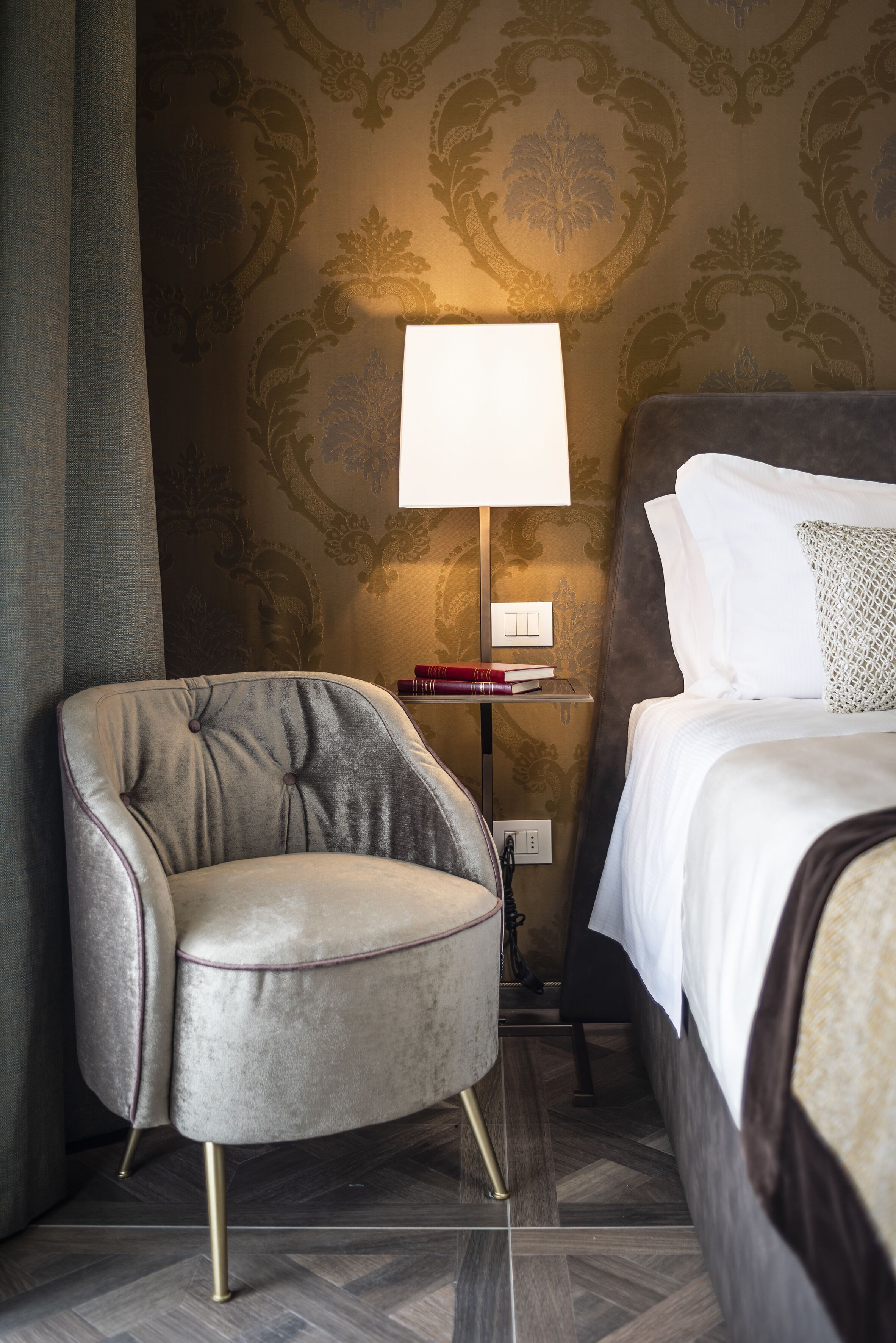 A hotel room with a tufted armchair, a bedside table with books, a lamp, and a neatly made bed.