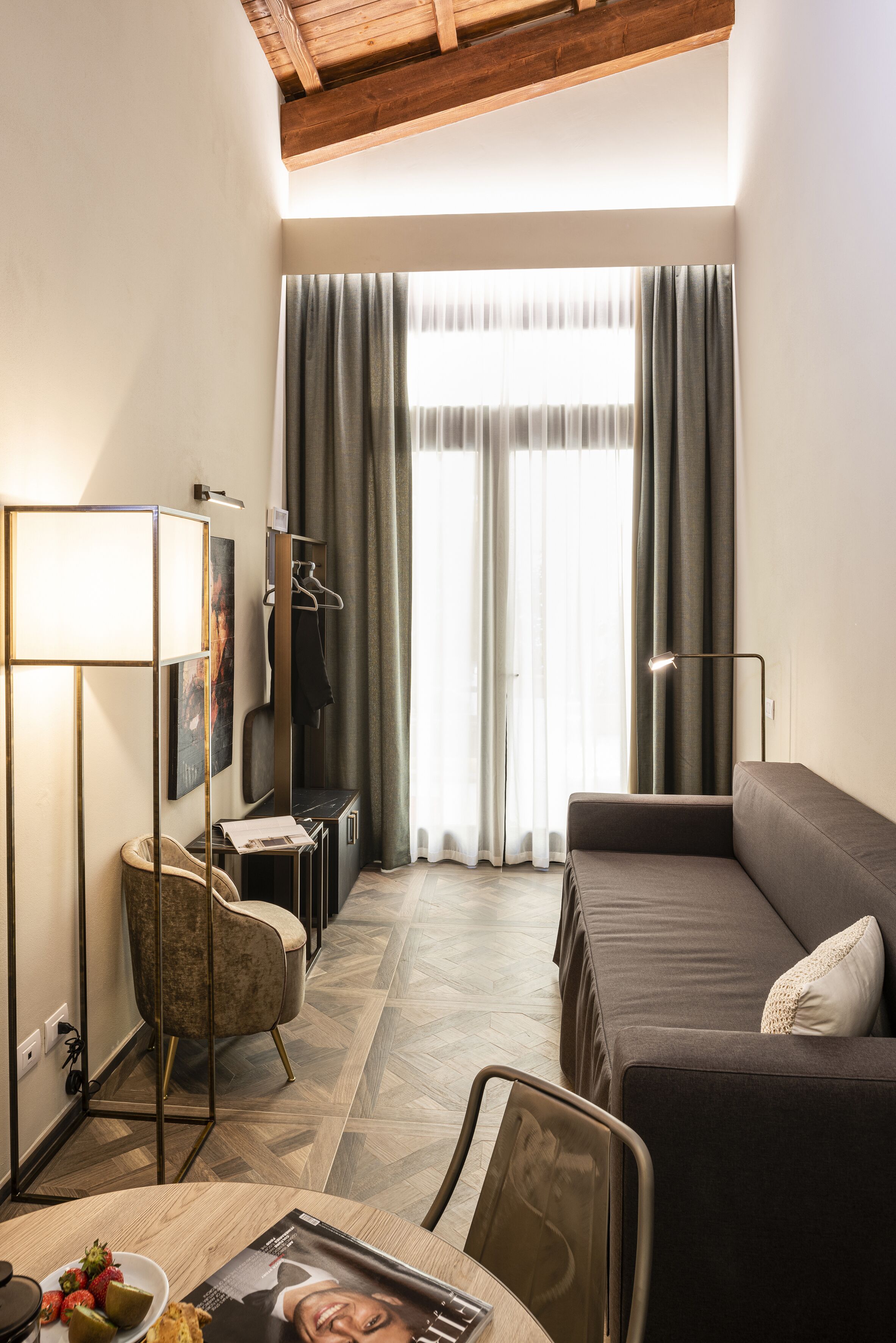 A modern hotel room with a sofa, a chair, a table with a magazine and fruit, a lamp, and a large window with curtains.