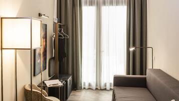A modern hotel room with a sofa, a chair, a table with a magazine and fruit, a lamp, and a large window with curtains.