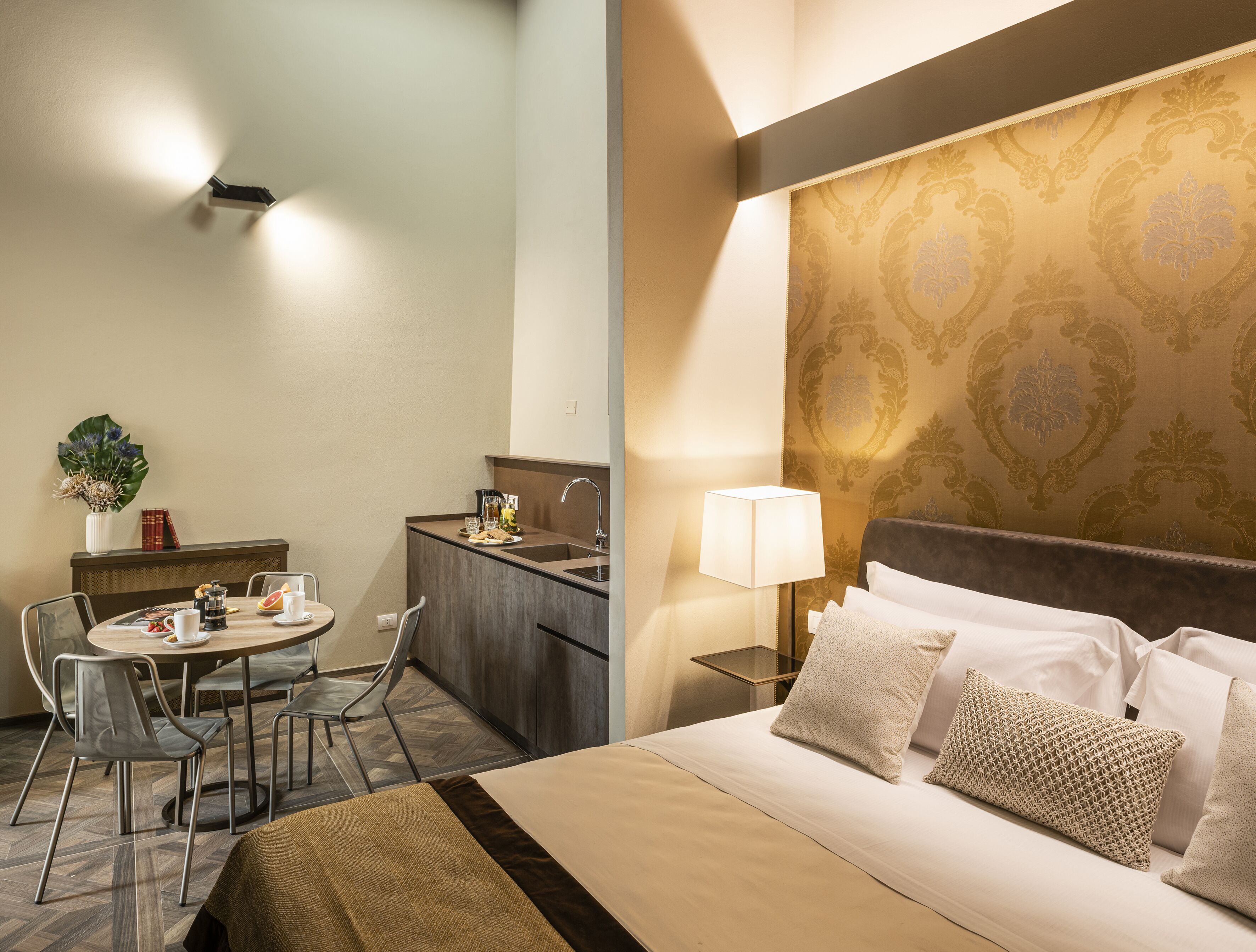 A hotel room with a bed, bedside table, lamp, and a small kitchenette area.