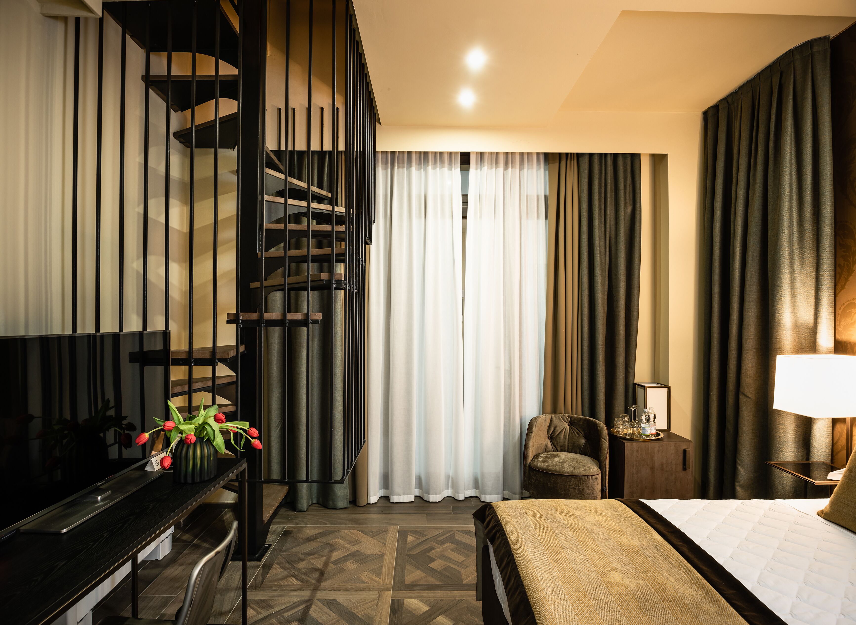 A modern hotel room with a bed, a desk, a chair, a small table, a lamp, and a staircase.