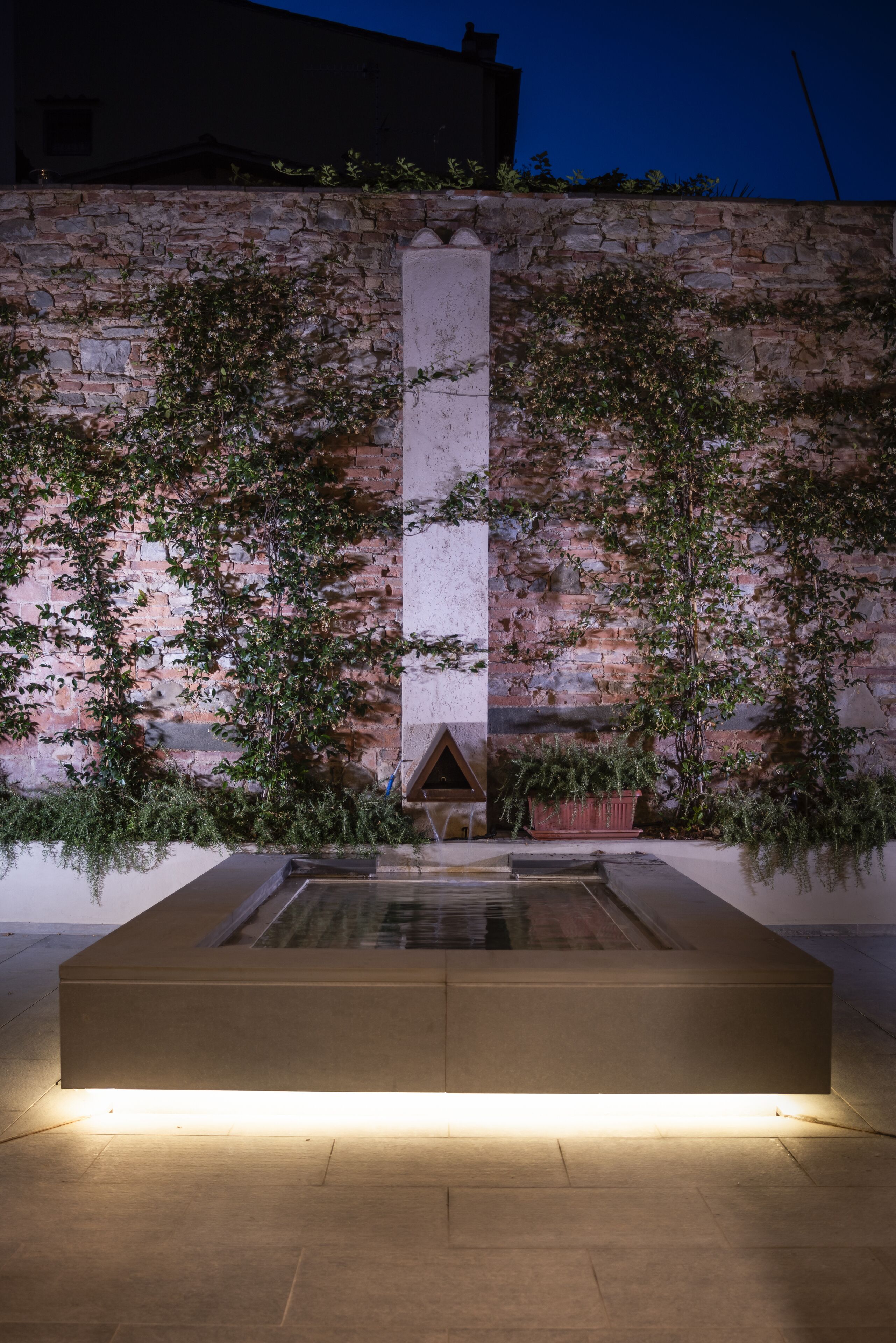 Un muro de piedra con plantas trepadoras, una fuente rectangular de agua y un banco de concreto.