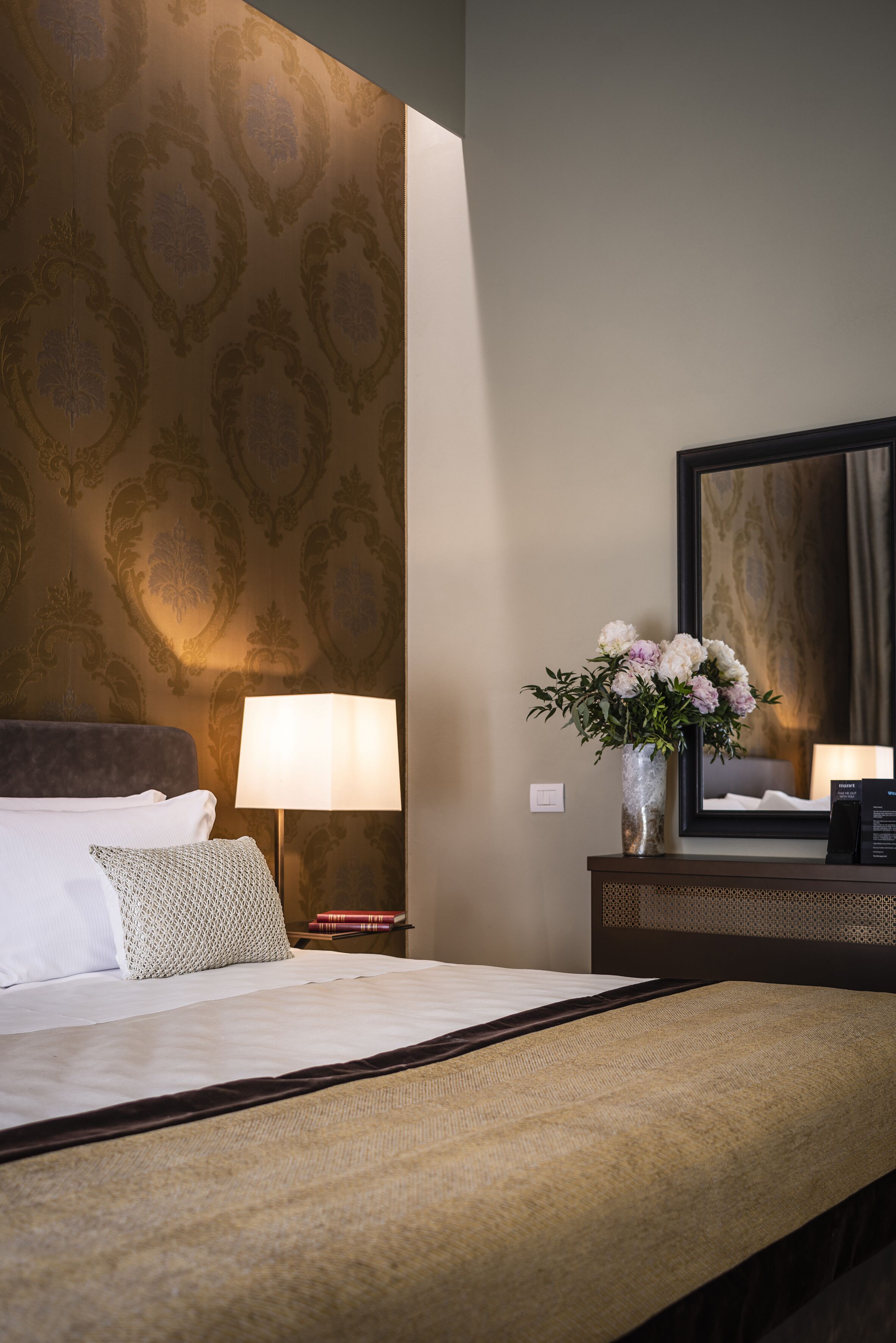 Habitación de hotel con cama, mesita de noche con lámpara, un jarrón con flores y un espejo en la pared.