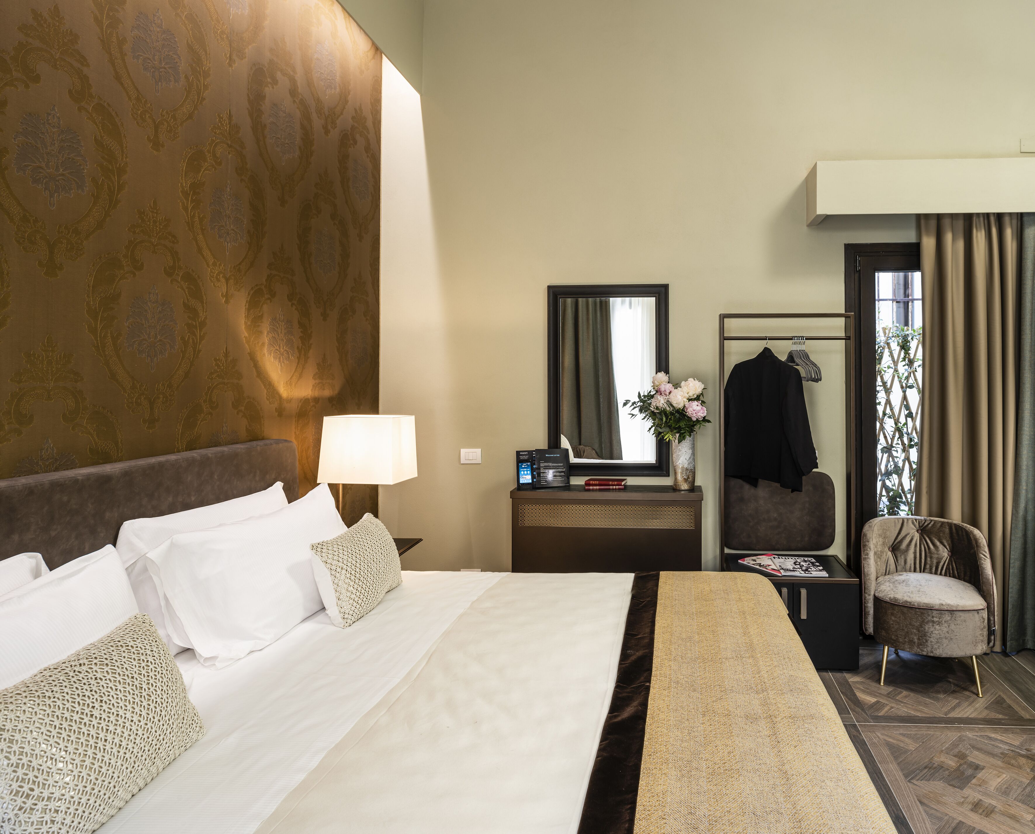 Habitación de hotel con una cama grande, una mesita de noche con lámpara, un espejo, una silla y una ventana con cortinas.