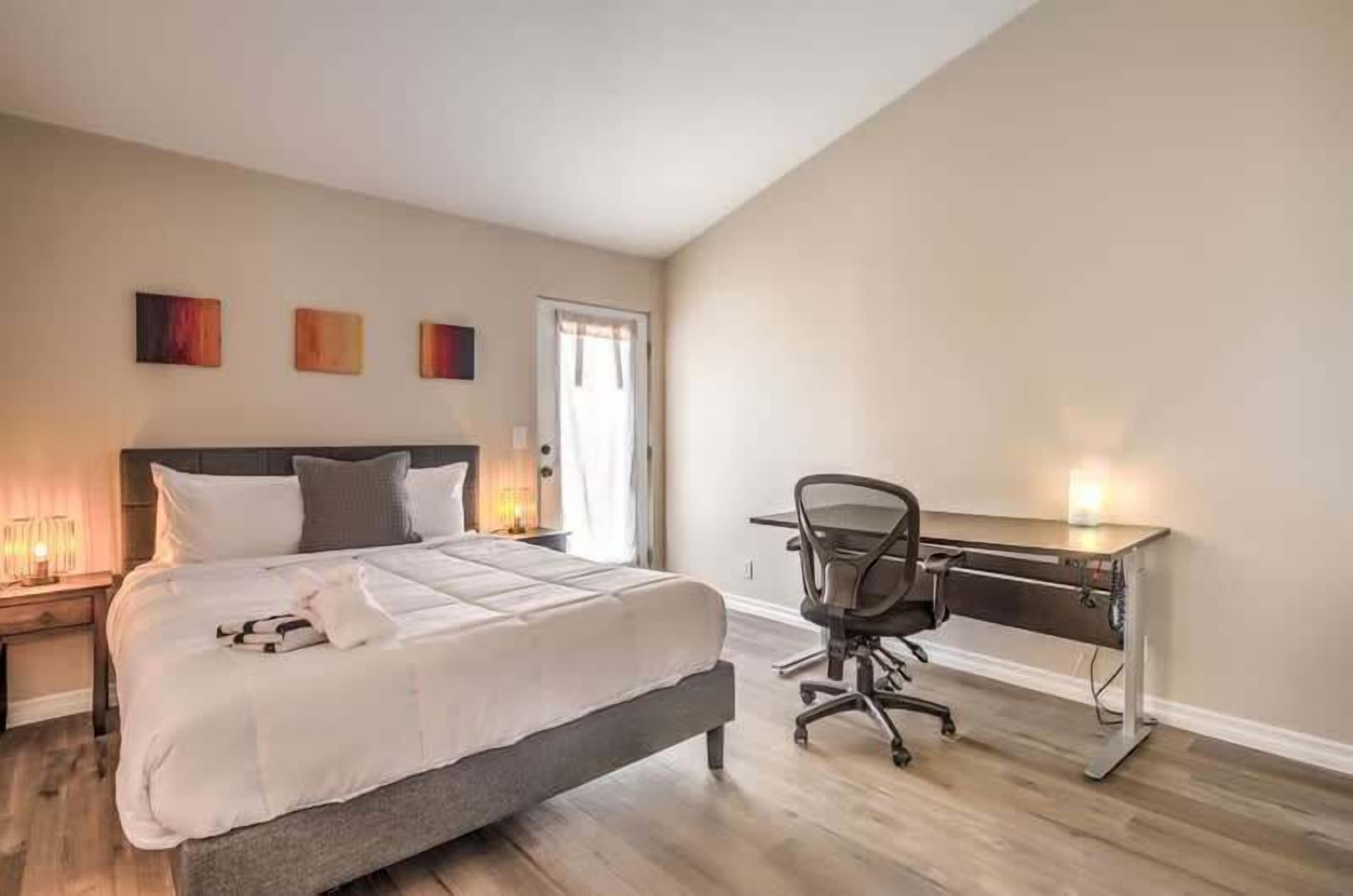 A bedroom with a bed, bedside table, lamp, desk, chair, and artwork on the wall.