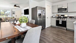 A modern kitchen with stainless steel appliances, a dining area with a wooden table and grey chairs, and a ceiling fan.