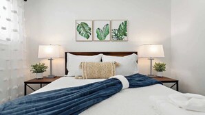 A neatly made bed with a blue blanket, white pillows, and a patterned throw. There are two bedside tables with lamps and small potted plants. Three framed botanical prints are hung on the wall above the bed.
