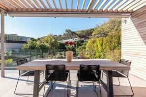 Eine Terrasse im Freien mit Holztisch und -stühlen, einem Blumenstrauß in einer Vase und Blick auf einen Hügel mit Bäumen und einem Gebäude im Hintergrund.